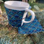 Bögremelegítő és egy alátét látszik a képen, egy fenyőágon fotózva. A kép egészen közeli, bal oldalon van a bögre, rajta a melegítő és jobb oldalon az alátét. A bögre kicsit az alátéten áll. A bögremelegítő és az alátét ugyanabból az anyagból készült, kék és fekete négyzetek, némi arannyal keretezve.