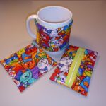 A képen egy szett látható, amely bögremelegítőből, bögrealátétből és zsebkendőtartóból áll. Mindegyik ugyanabból az anyagból készült, vidám, színes, kicsit gyerekrajzra hajaztó cicák tömegét ábrázolja. A bögre és a bögremelegítő van középen, bal oldalon az alátét, jobb oldalon a zsepitartó.