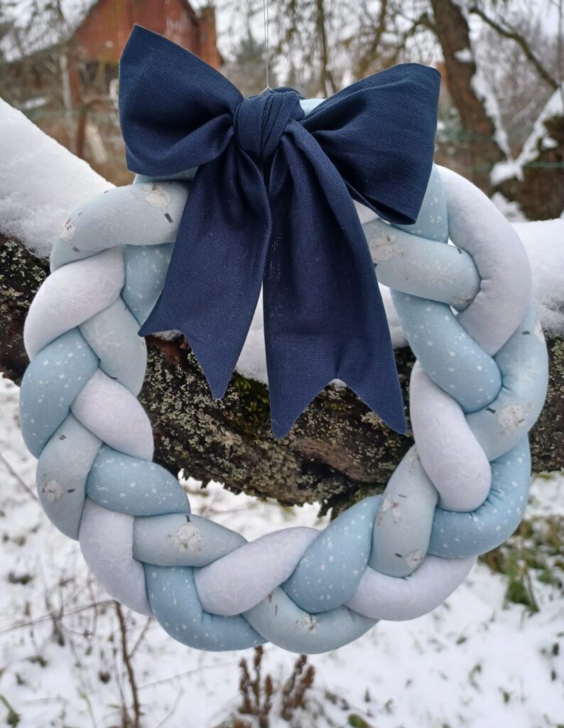 A képen egy textil koszorú látható, három féle anyagból van összefonva: fehér, világoskék és kék apró mintás. Egy hatalmas sötétkék koszorú díszíti a tetején. A háttérben egy havas faág látható.