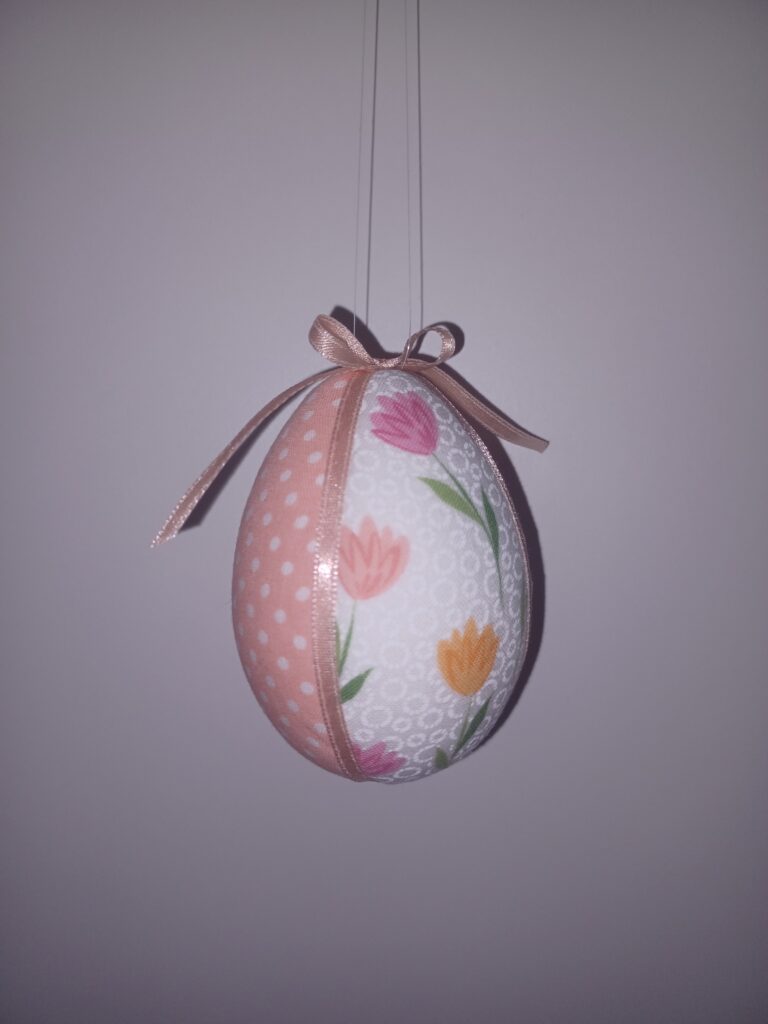 A képen egy húsvéti tojásdísz látható: hungarocell tojás, tavaszi textilekkel borítva, vékony szalaggal a tetején.